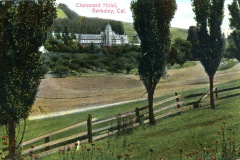 A scenic view of Claremont Hotel in Berkeley, California, surrounded by lush green hills and towering trees. A wooden fence runs across the foreground, with wildflowers dotting the grassy landscape.