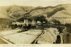 A vintage photograph of the Claremont Hotel in Berkeley, California. The hotel stands amidst hilly terrain with tennis courts in the foreground. The sepia tone adds an old-fashioned charm to the scenic landscape.