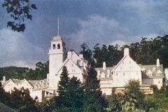 A large, historic white building with a central tower and steep roofs, surrounded by lush greenery and trees under a partly cloudy sky.