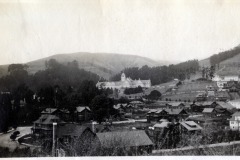 A vintage black and white photograph of a hillside town with a large, historic building in the center surrounded by trees. Smaller houses are scattered in the foreground, and rolling hills are visible in the background.
