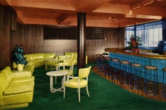 A retro-style lounge with lime green seating, a turquoise-tiled bar with round stools, and a green carpet. The room features wooden paneling, a central pillar, and a potted plant.