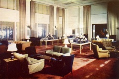 Vintage hotel lobby with high ceilings, large windows, and elegant decor. The space features plush armchairs, sofas, tables with floral arrangements, and soft lighting from scattered lamps, creating a warm, inviting atmosphere.