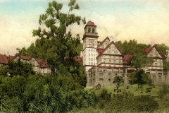 A vintage illustration of a large, historic building with multiple levels and red rooftops, surrounded by lush green trees and bushes. The building features a prominent tower, and the sky is a gradient of blue and pale peach.