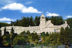 A grand, historic hotel surrounded by lush greenery and tall trees under a clear blue sky with a few clouds. The building has a central tower and multiple gabled roofs. Palm trees and well-kept gardens are in the foreground.