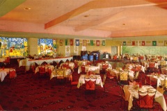 A retro-style restaurant with red carpet and tables set with white tablecloths. Vibrant stained glass windows decorate the back wall. Colorful banners hang above a buffet area in a spacious dining room.
