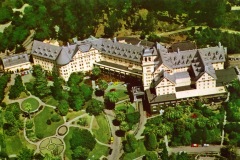 Aerial view of a sprawling, historic hotel surrounded by lush gardens and trees. The building is composed of multiple wings with peaked roofs and numerous windows. Pathways wind through the landscaped grounds, dotted with circular flower beds.