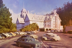 A large historic hotel with a tower stands amidst trees under a clear sky. A parking lot in the foreground is filled with vintage cars from the mid-20th century. The scene is bathed in warm sunlight.