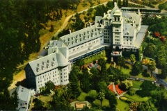 Aerial view of a large, historic hotel surrounded by lush greenery. The building has a white facade, multiple peaked roofs, and a central tower. Gardens with vibrant flowerbeds and palm trees are visible in the foreground.