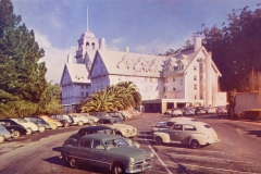 A vintage hotel with a tall central tower and large chimneys is surrounded by parked classic cars. The building is set against a background of trees under a clear sky. The scene evokes a nostalgic mid-20th-century feel.
