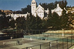 A scenic outdoor tennis court featuring several players in action. Beyond the court, a large, historic building with a tower is partially surrounded by trees under a clear blue sky.