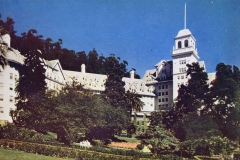 A grand, multi-story building with a prominent tower, surrounded by lush trees and a manicured garden under a clear blue sky. The architecture features gabled roofs and multiple windows.