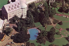 Aerial view of a large, historic hotel with white roof surrounded by lush greenery. A blue swimming pool and a patio area are visible on the grounds. Pathways wind through landscaped gardens with tall trees.