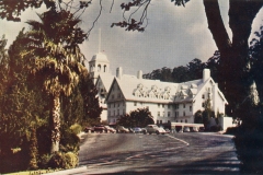 A large historic hotel with a white exterior and a central clock tower set amidst trees. Several cars are parked in front, and a palm tree is visible on the left. The sky is overcast.