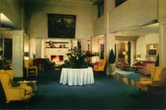 Spacious, elegant hotel lobby with high ceilings, featuring a central round table with a white tablecloth and plant centerpiece. Surrounded by plush seating, a fireplace, and warm lighting, evoking a cozy, inviting atmosphere.