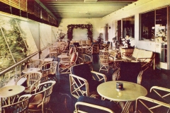 A vintage lounge with wicker chairs and round tables on a sunlit patio. Large windows on the left overlook lush greenery. The walls have neutral tones, and potted plants decorate the space, contributing to a relaxed, airy ambiance.