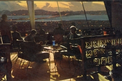A group of people sit at tables in a dimly lit, elevated restaurant or bar with large windows. The view outside shows a cityscape with twinkling lights and mountains against a sunset. Shadows are cast on the wooden floor.