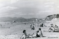 Black-and-white photo of people relaxing and sunbathing on a sandy San Francisco beach, with the iconic Cliff House perched on a cliff in the background and hills visible in the distance.