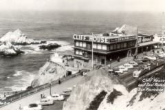 A historic black-and-white photo of Cliff House in San Francisco overlooks the ocean and Seal Rocks, with vintage cars parked along the road and waves crashing against the rocky coastline.
