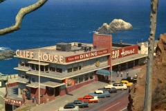 A vintage photo of Cliff House in San Francisco, a multi-story restaurant overlooking the ocean, with large signs and classic cars out front. The rocky coastline and sea complete this nostalgic scene.