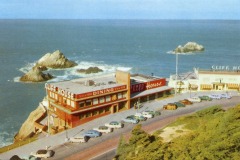 A vintage postcard view of San Francisco’s Cliff House, a seaside restaurant with red and yellow signage, overlooking rocky shores and the ocean. Several classic cars are parked along the road in front of the building.
