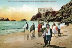 Children and adults in early 20th-century clothing stand on a sandy beach near the ocean, with rocky cliffs and San Francisco’s historic Cliff House building in the background.