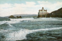 A historic photograph of the Cliff House, a grand Victorian landmark in San Francisco, perched on a rocky bluff above the Pacific Ocean with waves crashing below and hills rising in the background.