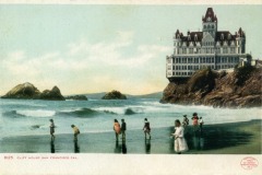 People wade along the shore near the historic Cliff House in San Francisco, a large, ornate building perched on a rocky bluff above the ocean with waves crashing nearby.