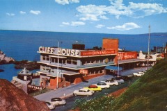 A vintage postcard shows San Francisco’s iconic Cliff House restaurant perched on a seaside cliff, with classic cars parked out front and the ocean and rocky shoreline stretching beneath a blue sky with scattered clouds.