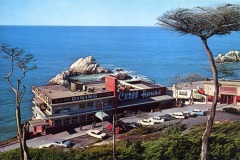 A vintage view of San Francisco’s iconic Cliff House restaurant overlooking the ocean and rocky shoreline, with classic cars parked in front and windswept trees framing the scene under a clear blue sky.