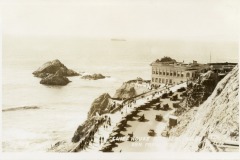 Historic photo of Cliff House in San Francisco, perched on a cliff above the ocean, with waves crashing on rocks below, cars parked along a curved road, and a ship visible in the distant water.