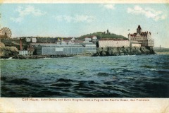 Vintage postcard showing Cliff House and Sutro Baths on San Francisco’s rocky coastline, viewed from the ocean. Large buildings and a Ferris wheel sit atop and near the cliffs above the crashing waves.