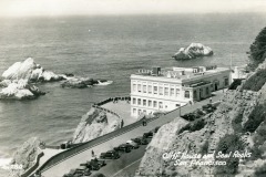 Black-and-white photo of the historic Cliff House in San Francisco, perched above the ocean with parked cars and rocky outcrops behind. Text reads: “Cliff House at Seal Rocks, San Francisco.”.