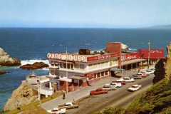 A vintage photo of the Cliff House in San Francisco, perched above the ocean with waves crashing on rocks below. Several cars line the street, and a gift shop stands beside the iconic restaurant.