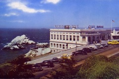 Historic postcard image of San Francisco’s iconic Cliff House restaurant perched on a hill overlooking the ocean, with several vintage cars parked along the road and rugged rocks jutting out of the water nearby.