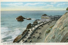 A vintage postcard features Cliff House by the ocean in San Francisco, with early automobiles parked along a curving coastal road and rocky outcrops emerging from the sea.