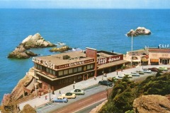 A vintage photo of the Cliff House in San Francisco, overlooking the ocean with parked cars along the road, rocky outcrops in the water, and a clear blue sky in the background.