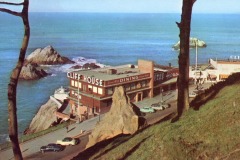 A vintage postcard view of Cliff House in San Francisco shows the iconic seaside restaurant perched on a rocky shoreline, with waves, parked cars, and people nearby, all seen from a grassy hill above.