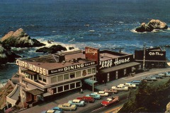 A vintage photo of the Cliff House restaurant in San Francisco, perched on a rocky coastline as waves crash below. Classic cars line the street, and a charming gift shop sits adjacent to the iconic main building.