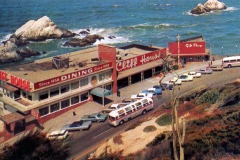 Aerial view of San Franciscos historic Cliff House restaurant and gift shop by the ocean, with parked cars and a tour bus along the coastal road; waves crash against rocks in the background.