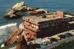 A vintage photo of Cliff House in San Francisco, a restaurant overlooking the ocean with waves, rocks, and classic cars parked in front. The building’s large sign and iconic mid-century architecture are visible.