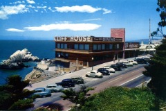 A vintage photo of San Francisco’s iconic Cliff House restaurant overlooking the ocean, with classic 1940s cars parked along the road and blue sky above. The coastline and large rocks are visible in the background.