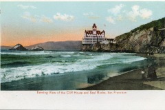 A vintage postcard shows San Franciscos ornate Cliff House perched on a rocky bluff, overlooking ocean waves and Seal Rocks at sunset. Two people stand on the sandy shore in the foreground.