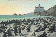 A large crowd of people in early 1900s clothing gathers on a sandy beach near the ocean, with waves, rocks, and San Francisco’s iconic Cliff House, a grand Victorian-style building, on a cliff in the background.
