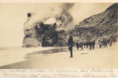 A group of people stand on a beach watching San Francisco’s Cliff House on a rocky cliff engulfed in smoke, with handwritten text in German at the bottom of the sepia-toned photograph.