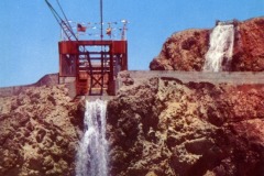 A cable car tower sits between rocky cliffs, with a waterfall flowing down each side. A white fence and part of a bridge are visible below under a clear blue sky near San Francisco’s famous Cliff House.