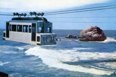 A cable car suspended above ocean waves travels toward a large rocky outcrop near San Franciscos famous Cliff House, with blue sky and water in the background.