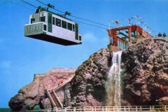 A cable car rides high above a rocky cliff with a waterfall, approaching the bright red Cliff House station in San Francisco, adorned with flags under a blue sky. People are visible on the stairs and near the station.