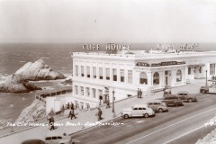 Historic black-and-white photo of San Francisco’s iconic Cliff House restaurant on Ocean Beach, with vintage cars parked outside and waves crashing against rocks in the background.