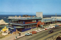 A vintage seaside street scene in San Francisco with colorful cars parked along a road near the iconic Cliff House, a blue building adorned with an ocean wave mural, overlooking rocky cliffs and the sparkling ocean beneath a clear sky.