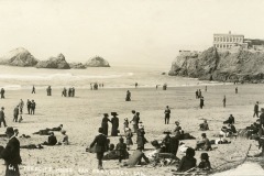 Black-and-white photo of people in early 1900s clothing walking and lounging on a San Francisco beach, with large rocks in the ocean and the iconic Cliff House perched on a cliff in the background.